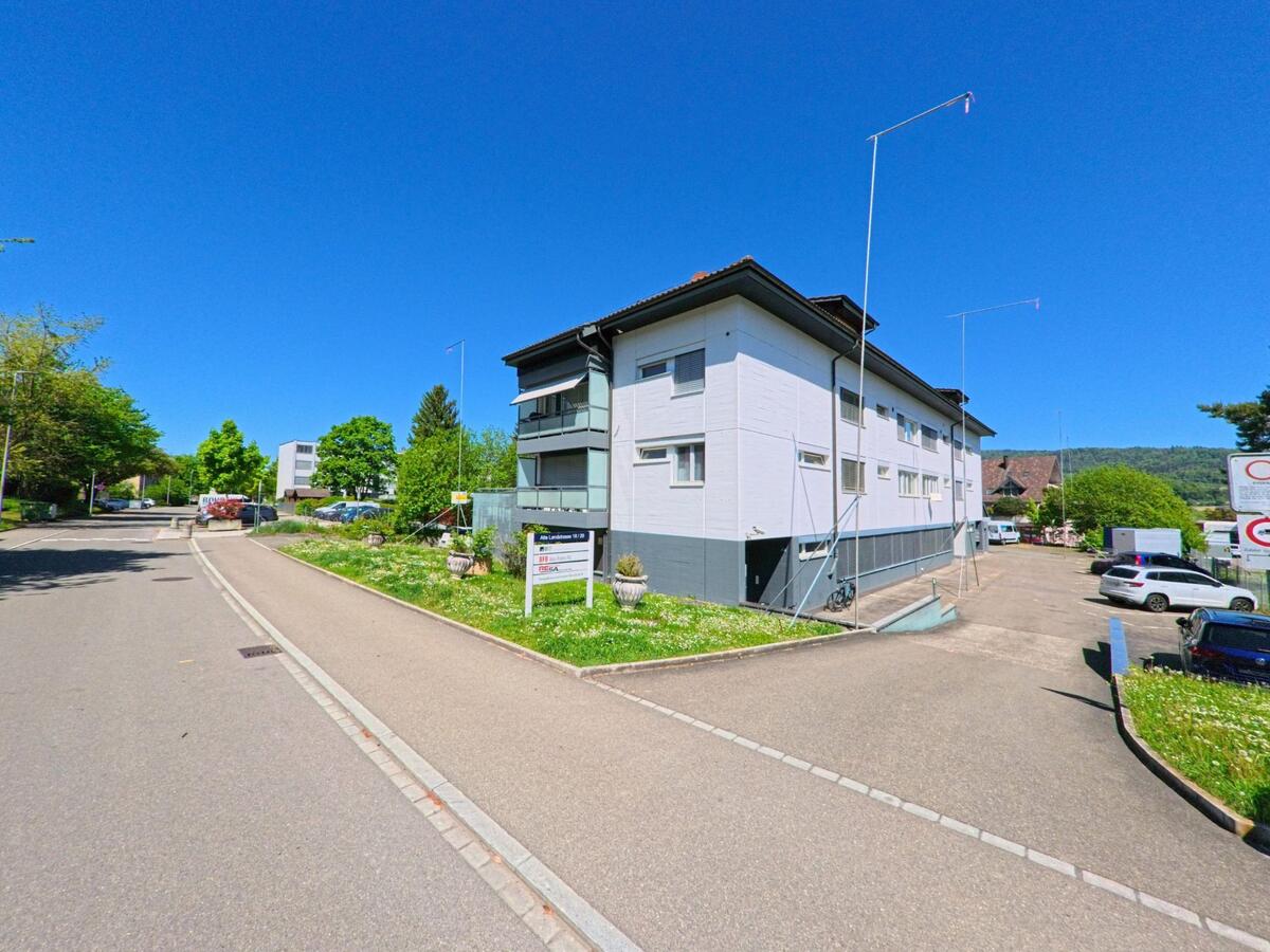 Ein Mehrfamilienhaus mit Parkplätzen und grüner Umgebung steht in der Gemeinde Dänikon im Kanton Zürich, PLZ 8114, nahe einer ruhigen Seitenstrasse und mit Blick auf bewaldete Hügel.
