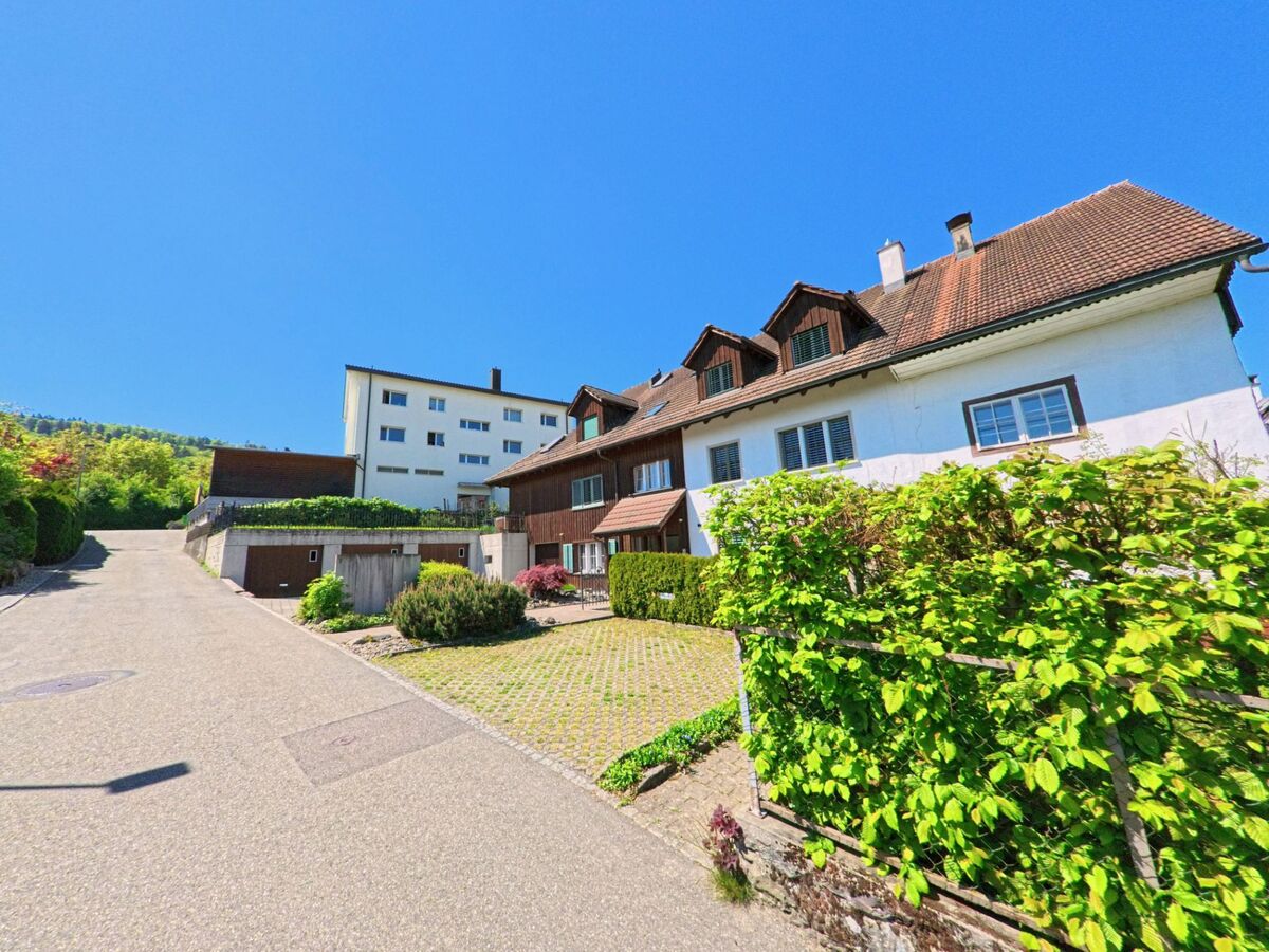 In Dänikon im Kanton Zürich, PLZ 8114, führt eine ruhige Seitenstrasse an gepflegten Wohnhäusern vorbei, die sich an einem sanften Hang mit Blick auf bewaldete Hügel befinden.