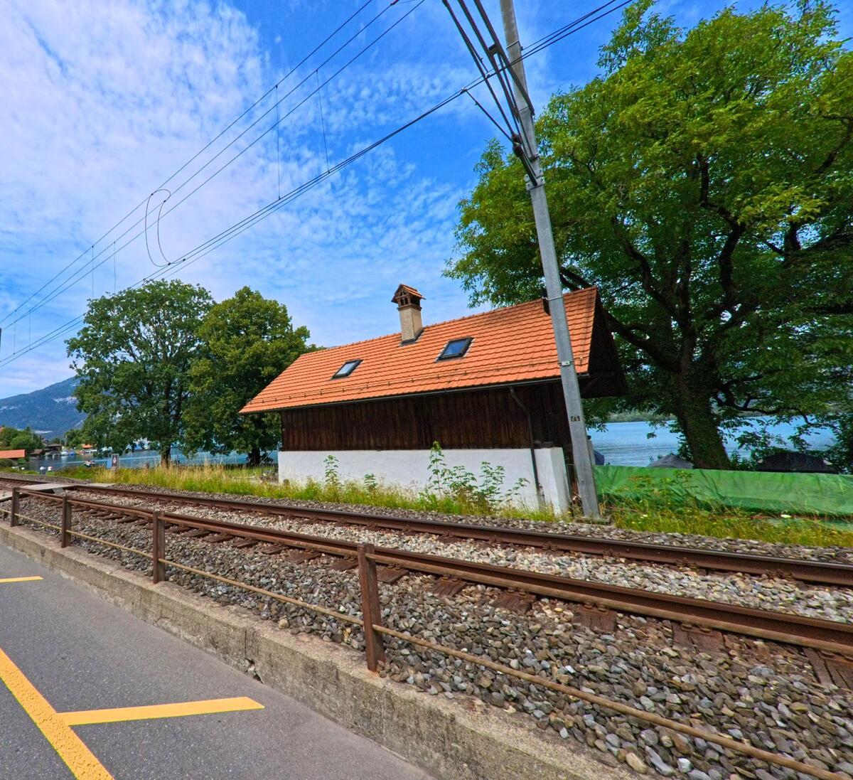 In Därligen (3707, Kanton Bern) verläuft ein Gleisabschnitt unmittelbar vor einem kleinen Gebäude mit Ziegeldach, während im Hintergrund der Thunersee zwischen Bäumen durchscheint.