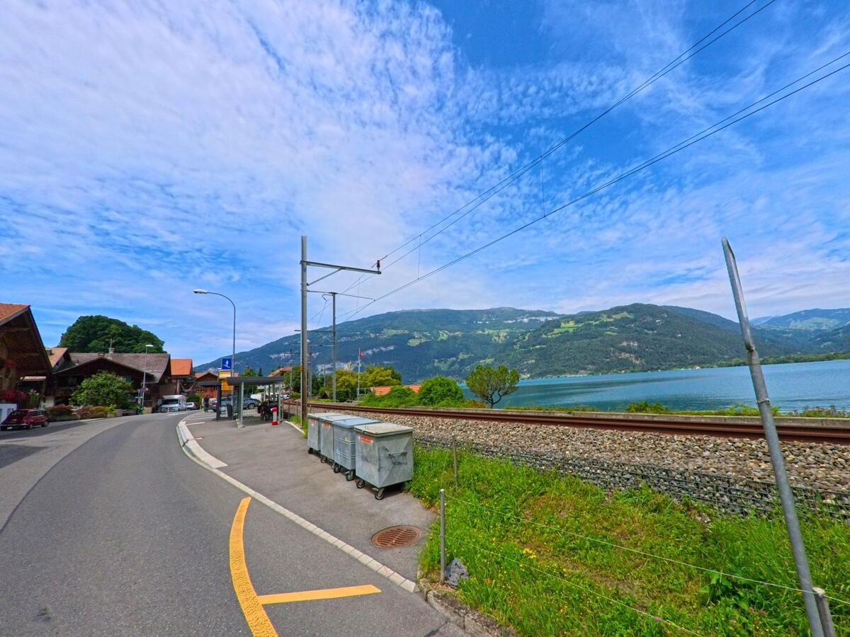 In Därligen (3707, Kanton Bern) verläuft eine schmale Strasse mit Bushaltestelle parallel zur Bahnlinie, die am Ufer des Thunersees entlangführt, mit Blick auf bewaldete Hänge.