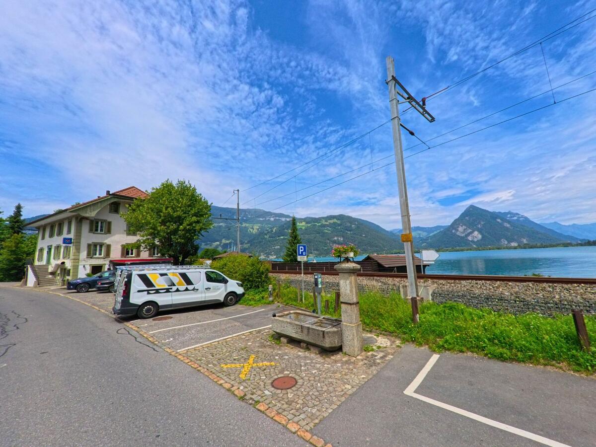 In Därligen (3707, Kanton Bern) steht ein historischer Brunnen aus Stein nahe der Bahnlinie, mit Blick auf den Thunersee und die umliegenden Berge im Hintergrund.