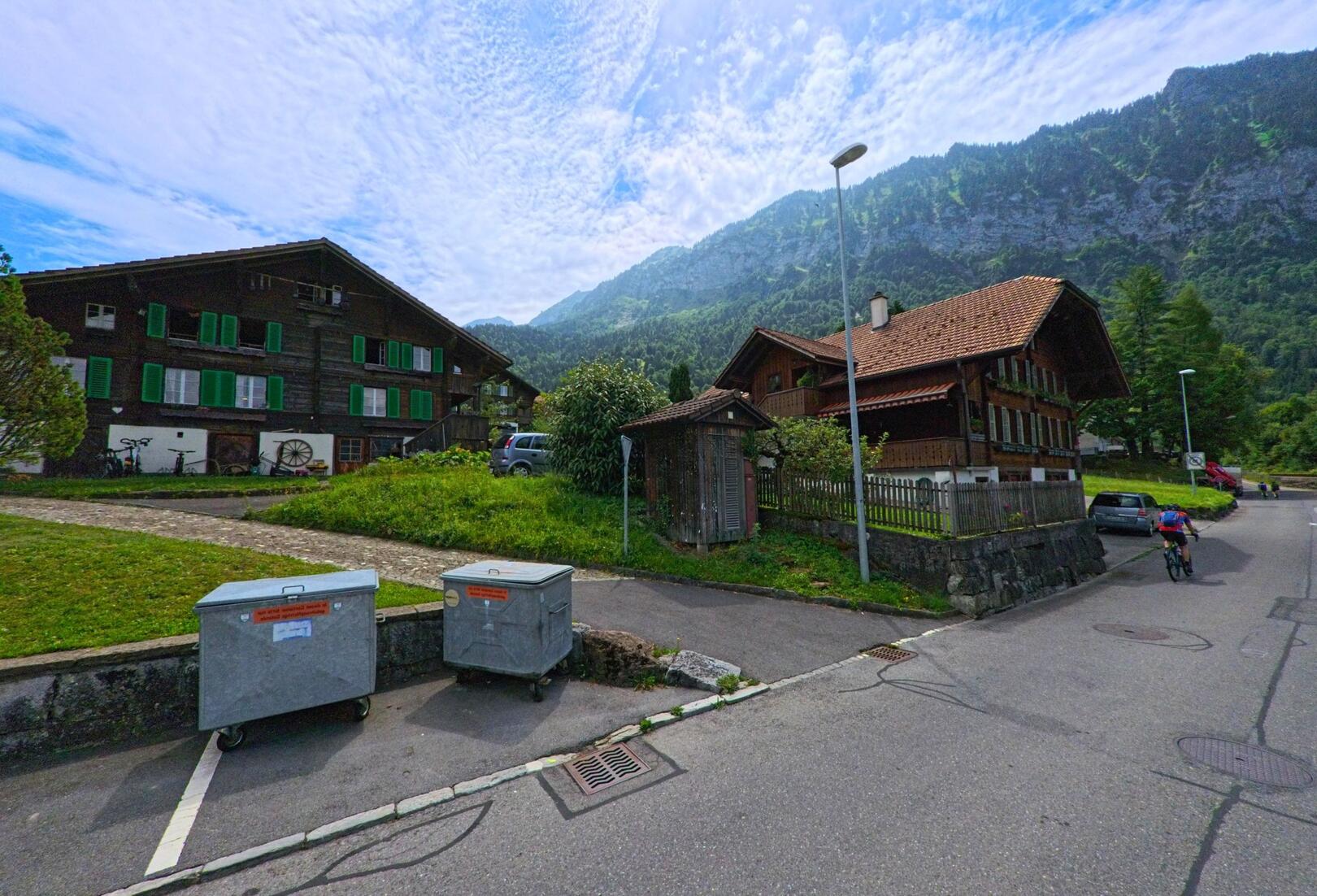 Im Zentrum von Därligen (3707, Kanton Bern) stehen traditionelle Holzhäuser mit grünen Fensterläden vor einer eindrucksvollen Bergkulisse, während ein Velofahrer die ruhige Strasse entlangfährt.