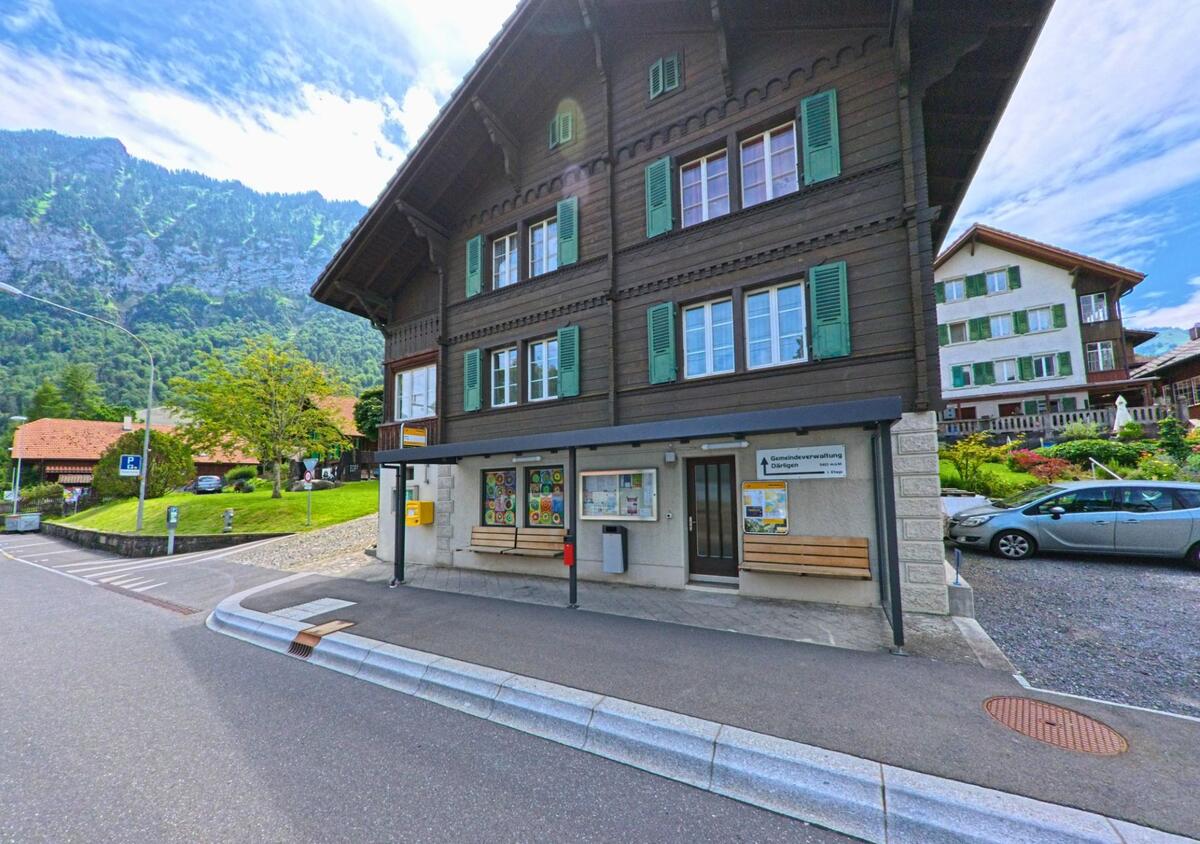 Vor der Gemeindeverwaltung von Därligen (3707, Kanton Bern) mit ihrem traditionellen Holzbau und grünen Fensterläden befindet sich eine überdachte Sitzbank mit Anschlagtafeln und Briefkasten.