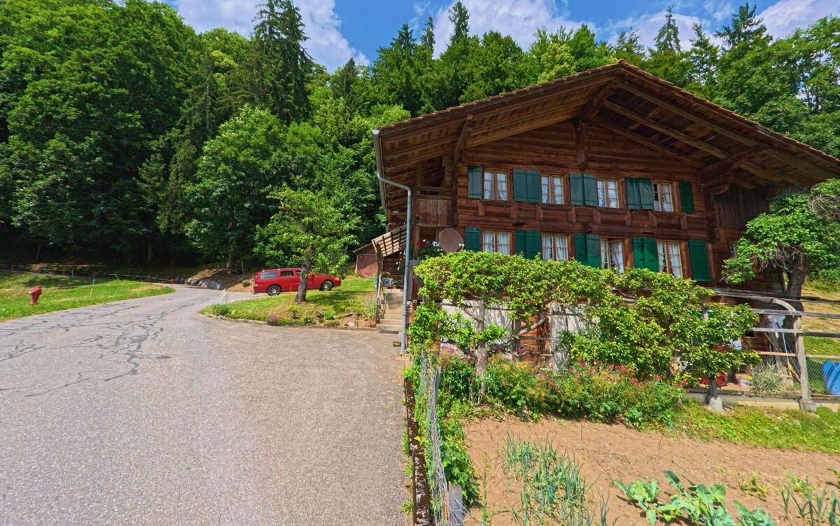 Direkt am Waldrand von Därstetten (3764, Kanton Bern) liegt ein traditionelles Holzhaus mit gruenen Fensterlaeden und kleinem Gemuesegarten an einer abzweigenden Strasse.