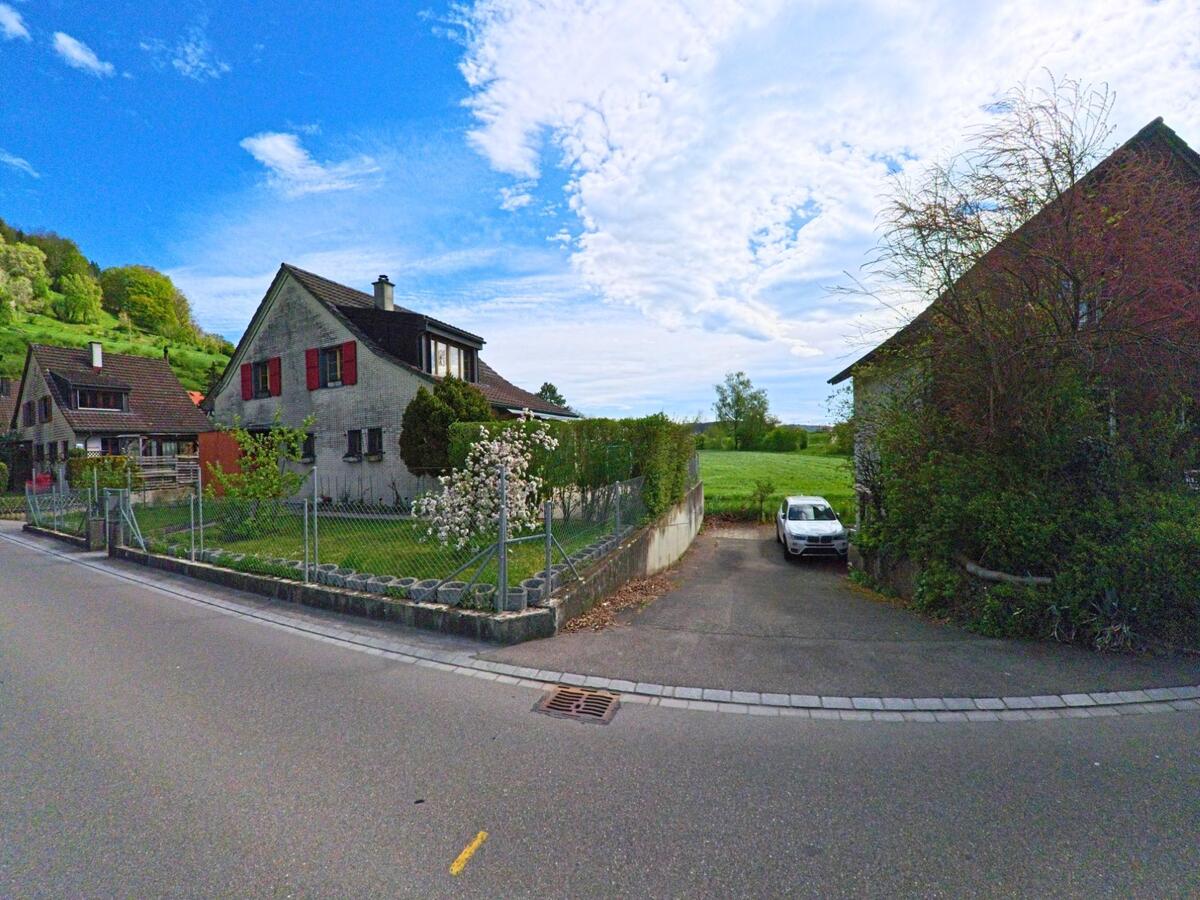 Ein Einfamilienhaus mit roten Fensterläden steht in Dättlikon im Kanton Zürich, PLZ 8421, an einer Landstrasse mit angrenzender Wiese und Obstbäumen.