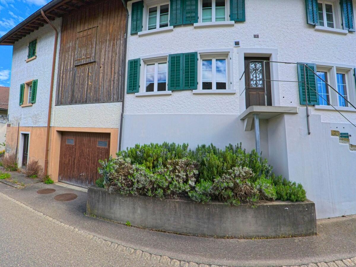 Ein traditionelles Wohnhaus mit grünen Fensterläden und Holzgarage steht in Dättlikon im Kanton Zürich, PLZ 8421, direkt an einer schmalen Strasse mit bepflanztem Vorbereich.