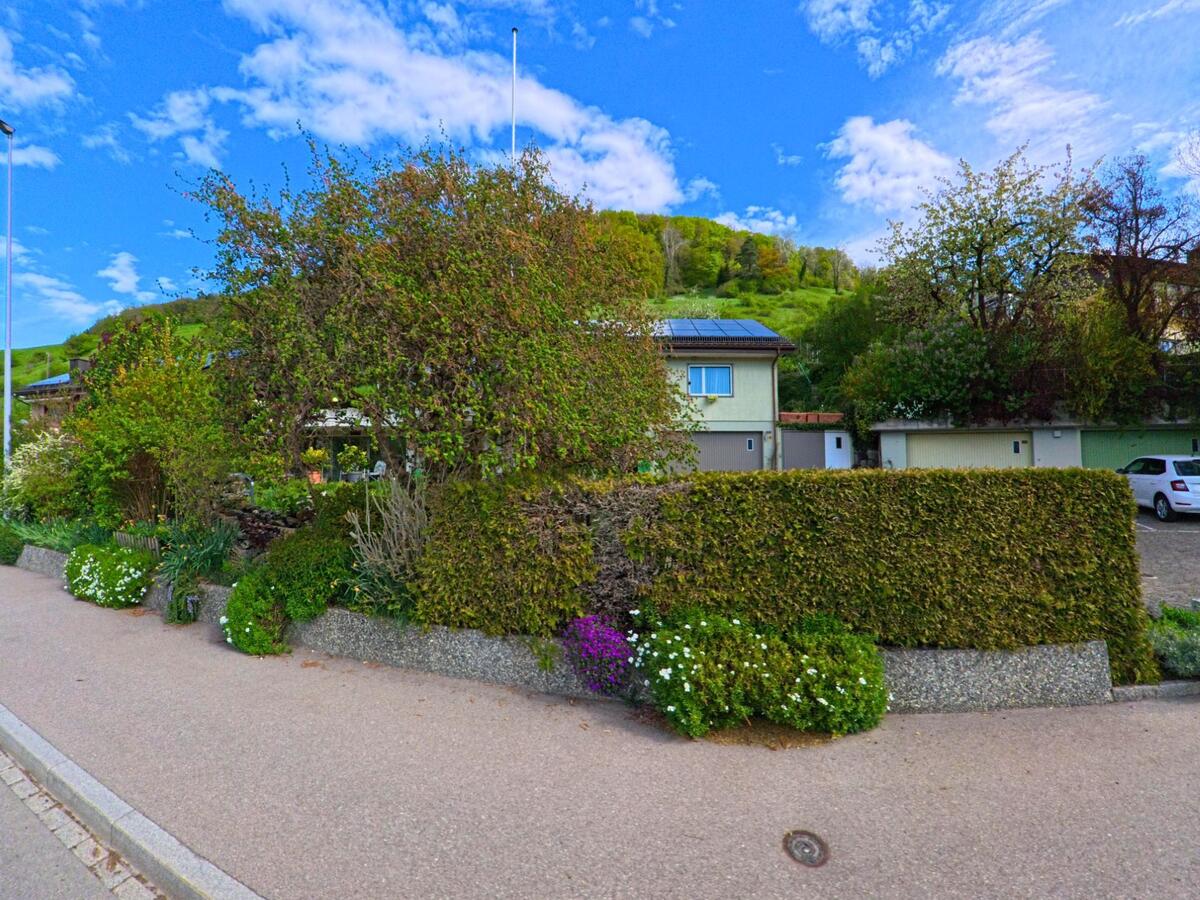 In Dättlikon im Kanton Zürich, PLZ 8421, verbirgt sich ein Wohnhaus mit Solarpanelen hinter dichter Bepflanzung und gepflegtem Vorgarten in einer ruhigen Wohngegend am Hang.