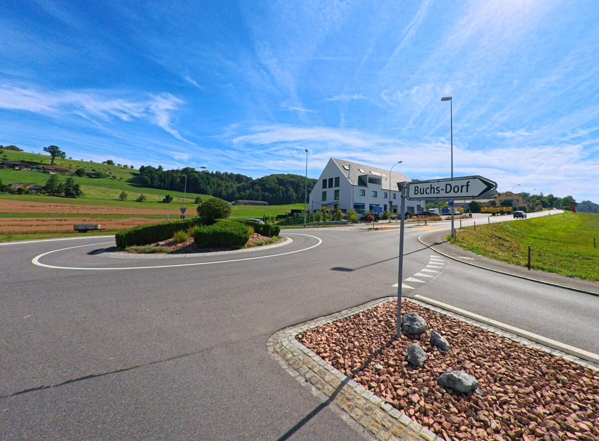  In Dagmersellen (6252, Kanton Luzern) fuehrt ein Verkehrskreisel an einer Wegweisung nach Buchs-Dorf vorbei, umgeben von Feldern und sanfter Huegellandschaft.