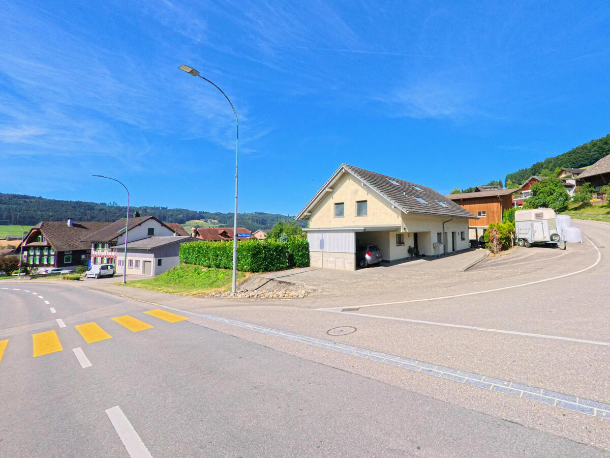 In Dagmersellen (6253, Kanton Luzern) trifft eine Nebenstrasse mit Bauernhaeusern und moderner Bebauung auf eine Hauptstrasse mit Fussgaengerstreifen und Blick auf die angrenzende Hügellandschaft.