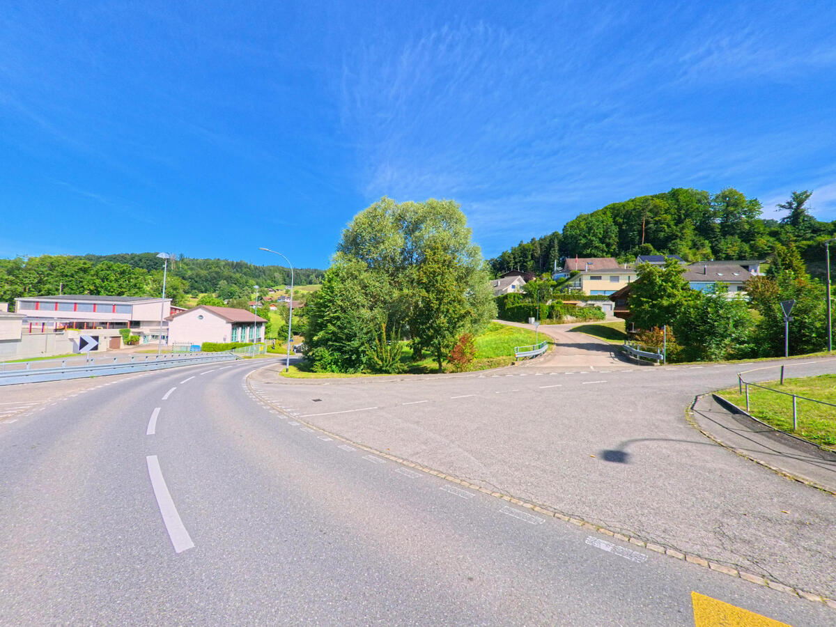 In Dagmersellen (6253, Kanton Luzern) verzweigt sich eine Strasse zwischen Hanglage und Siedlungsgebiet, flankiert von Baeumen, Einfamilienhaeusern und einem Schulgebaeude.
