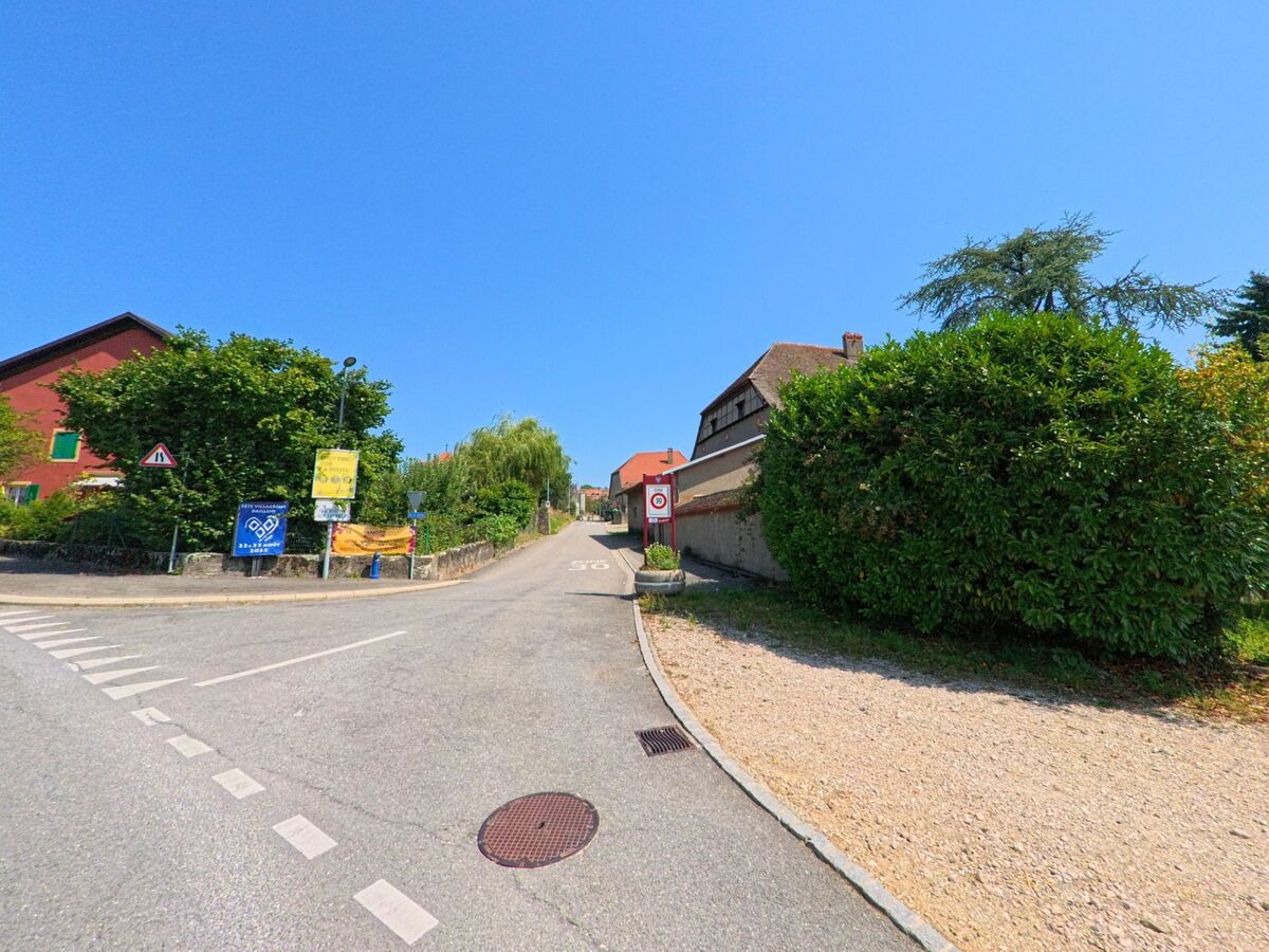 Eine schmale Strasse fuehrt zwischen Wohnhaeusern in Daillens (1306, Kanton Waadt) bergauf, gesaeumt von Hinweisschildern und dichter Vegetation.