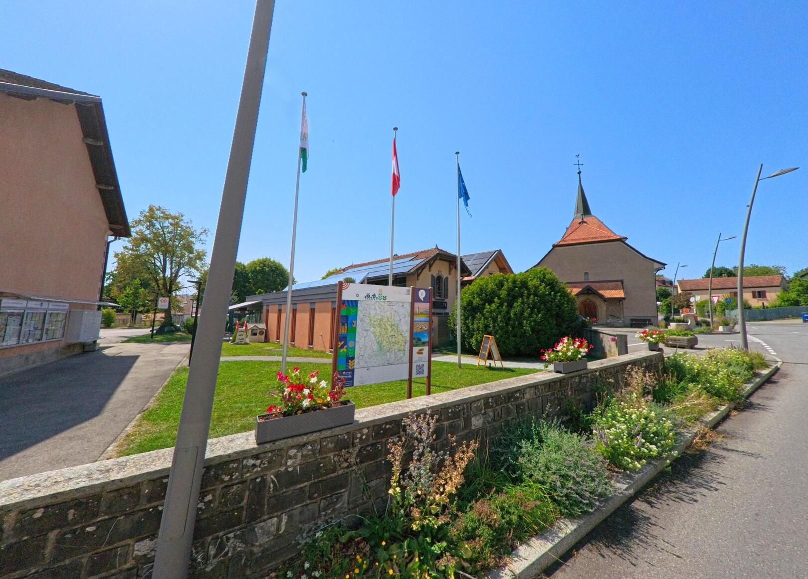 Vor der Kirche im Zentrum von Daillens (1306, Kanton Waadt) stehen drei Fahnenmasten und eine Infotafel, umgeben von gepflegten Blumenbeeten.