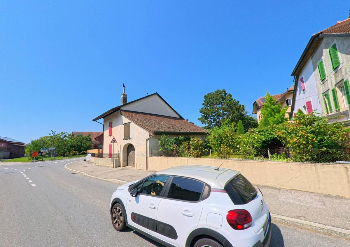 Ein kleines Haus mit roten Fensterlaeden steht an einer Strassenkurve in Daillens (1306, Kanton Waadt), waehrend ein Auto vorbeifaehrt.