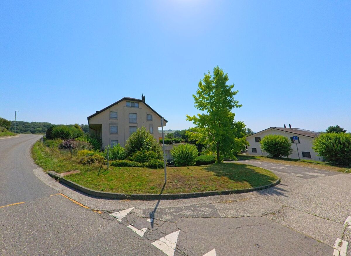 An einer kleinen Verkehrsinsel in Daillens (1306, Kanton Waadt) stehen ein Wohnhaus und ein einzelner Baum unter klarem Himmel.