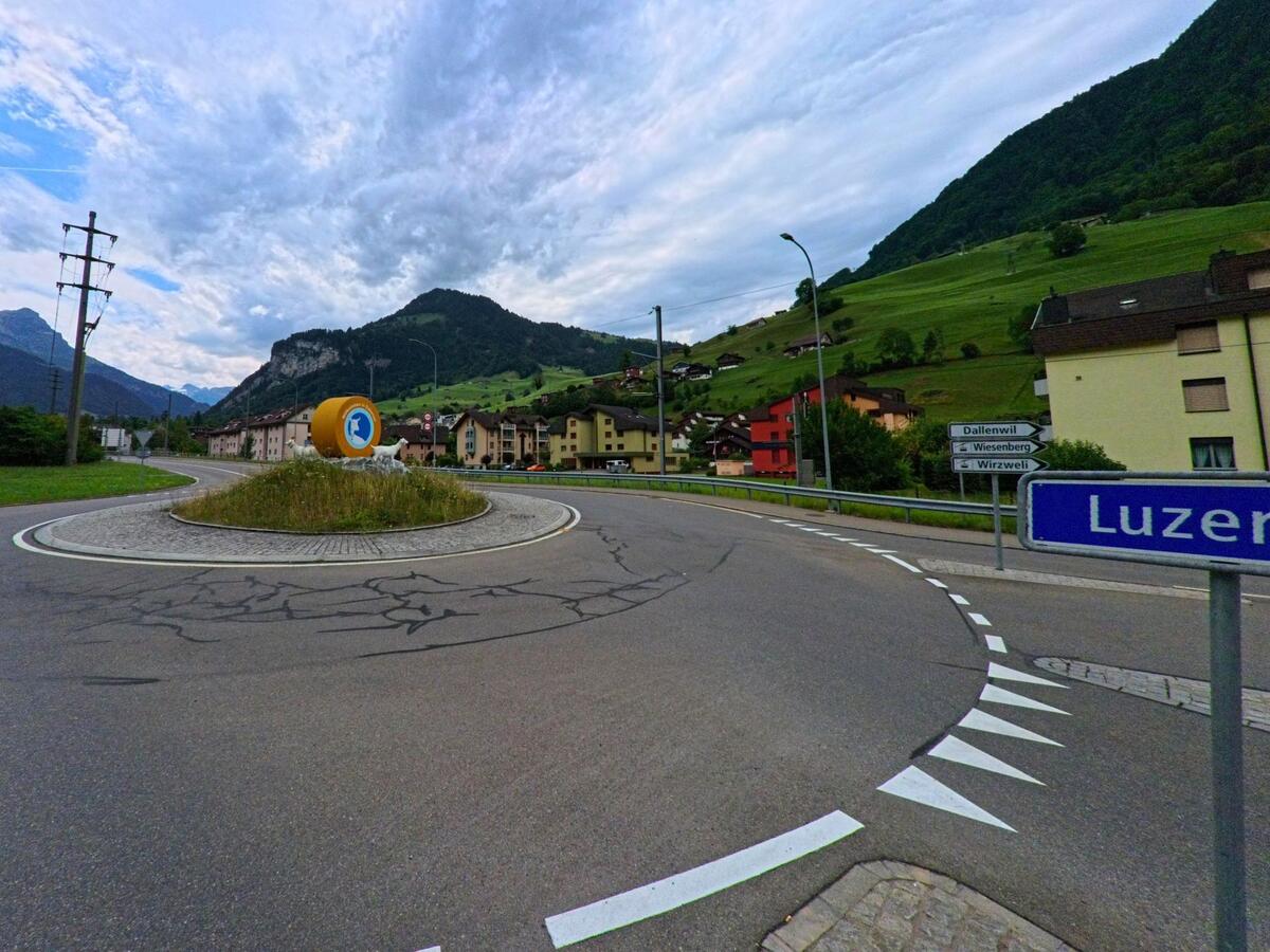 Am Ortseingang von Dallenwil (6382, Kanton Nidwalden) markiert ein Kreisverkehr mit auffaelliger Skulptur den Uebergang von Luzern in das Dorf, umgeben von gruener Hanglage und Wohnhaeusern.