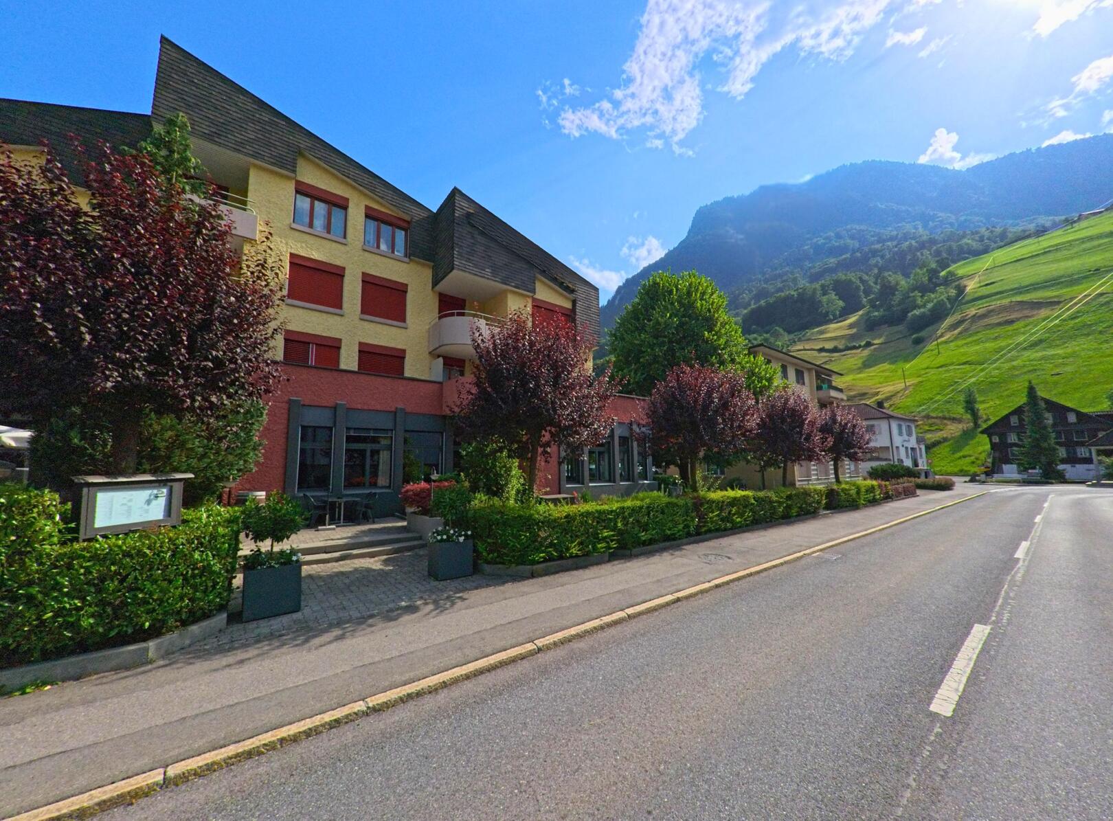 Entlang der Hauptstrasse von Dallenwil (6383, Kanton Nidwalden) steht ein modernes, mehrstöckiges Gebäude mit roten Fensterläden vor der Kulisse grüner Hänge und bewaldeter Berghänge.