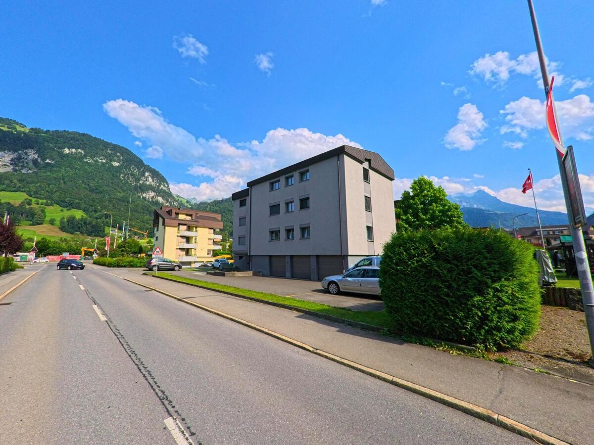 In der Gemeinde Dallenwil (6383, Kanton Nidwalden) verläuft eine breite Strasse an modernen Mehrfamilienhäusern vorbei, im Hintergrund ragen grüne Hänge und Berge empor.