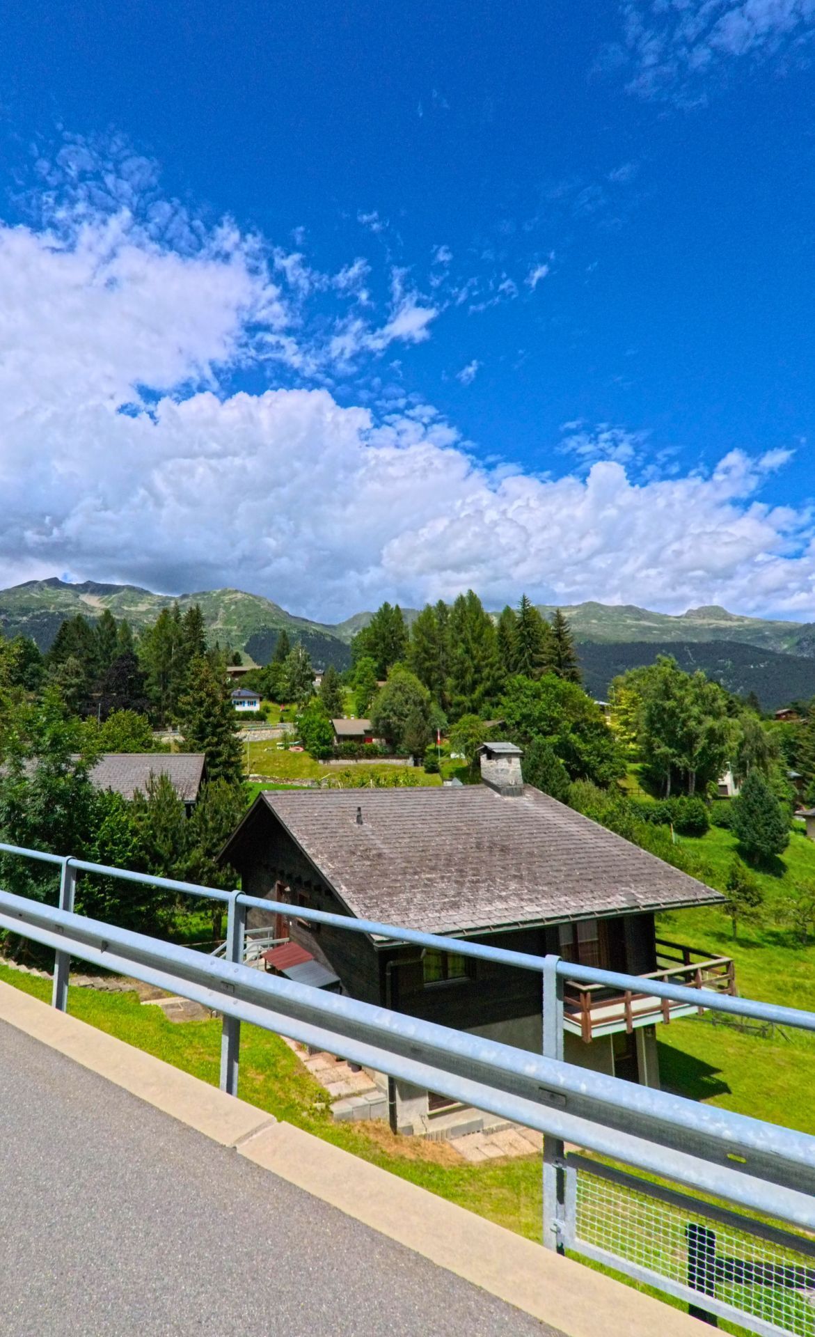 Ein Chalet mit Holzbalkon liegt in Dalpe (6774, Kanton Tessin) zwischen gruenen Baeumen und Wiesen, umgeben von den Bergen des Leventinatals.