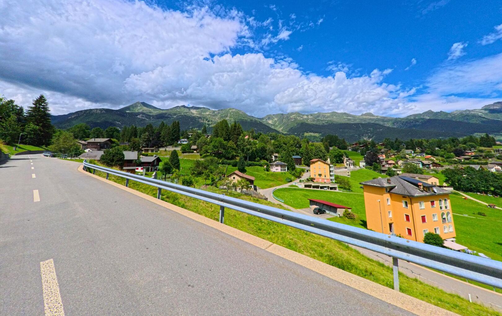Eine asphaltierte Strasse mit Geländer führt in Dalpe (6774, Kanton Tessin) an mehreren Wohnhäusern und grünen Wiesen vorbei, im Hintergrund erheben sich bewaldete Berghänge.