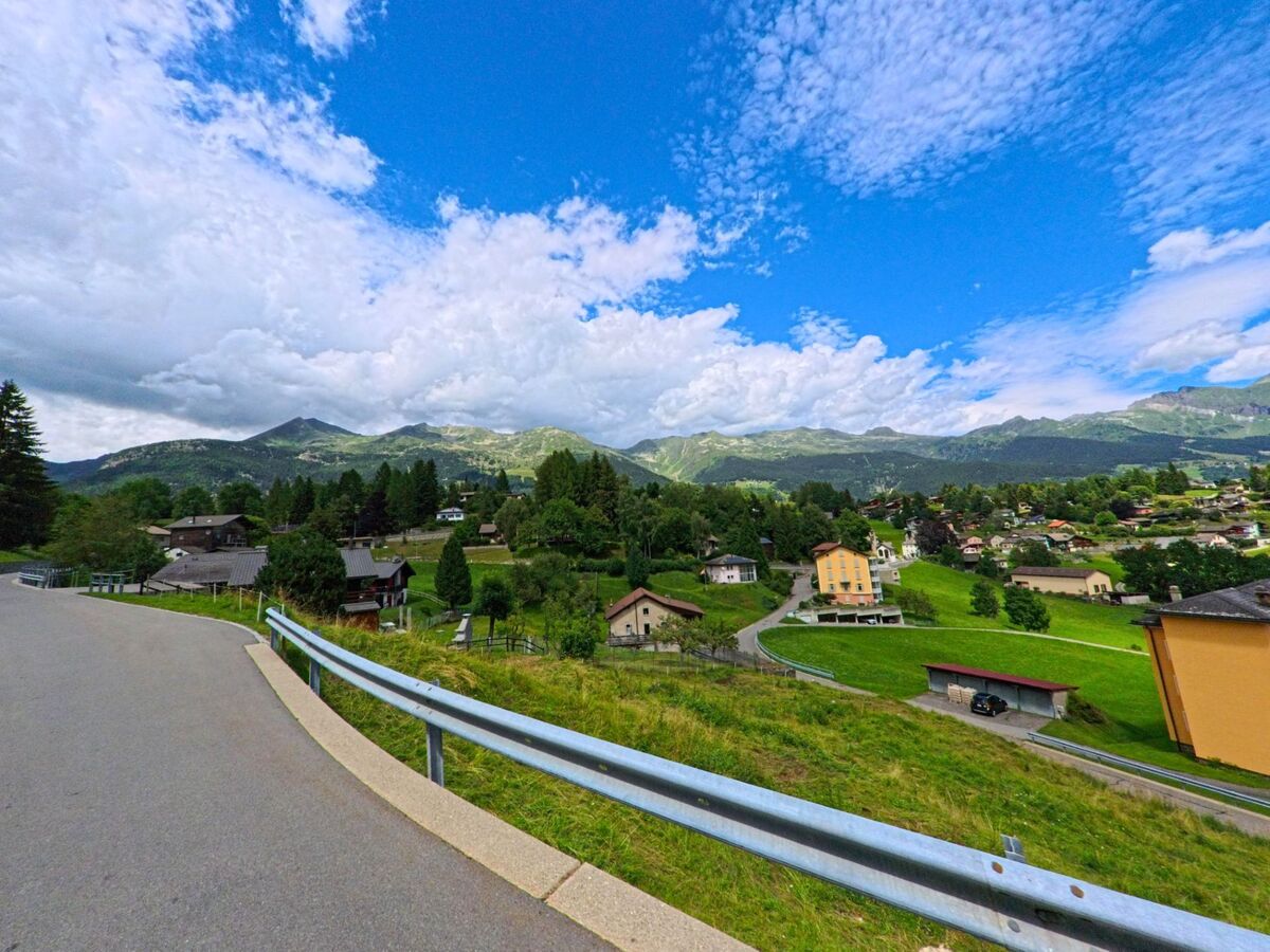 Eine kurvige Strasse mit Geländer fuehrt in Dalpe (6774, Kanton Tessin) durch ein Wohngebiet mit Wiesen und Blick auf die umliegenden Alpen.