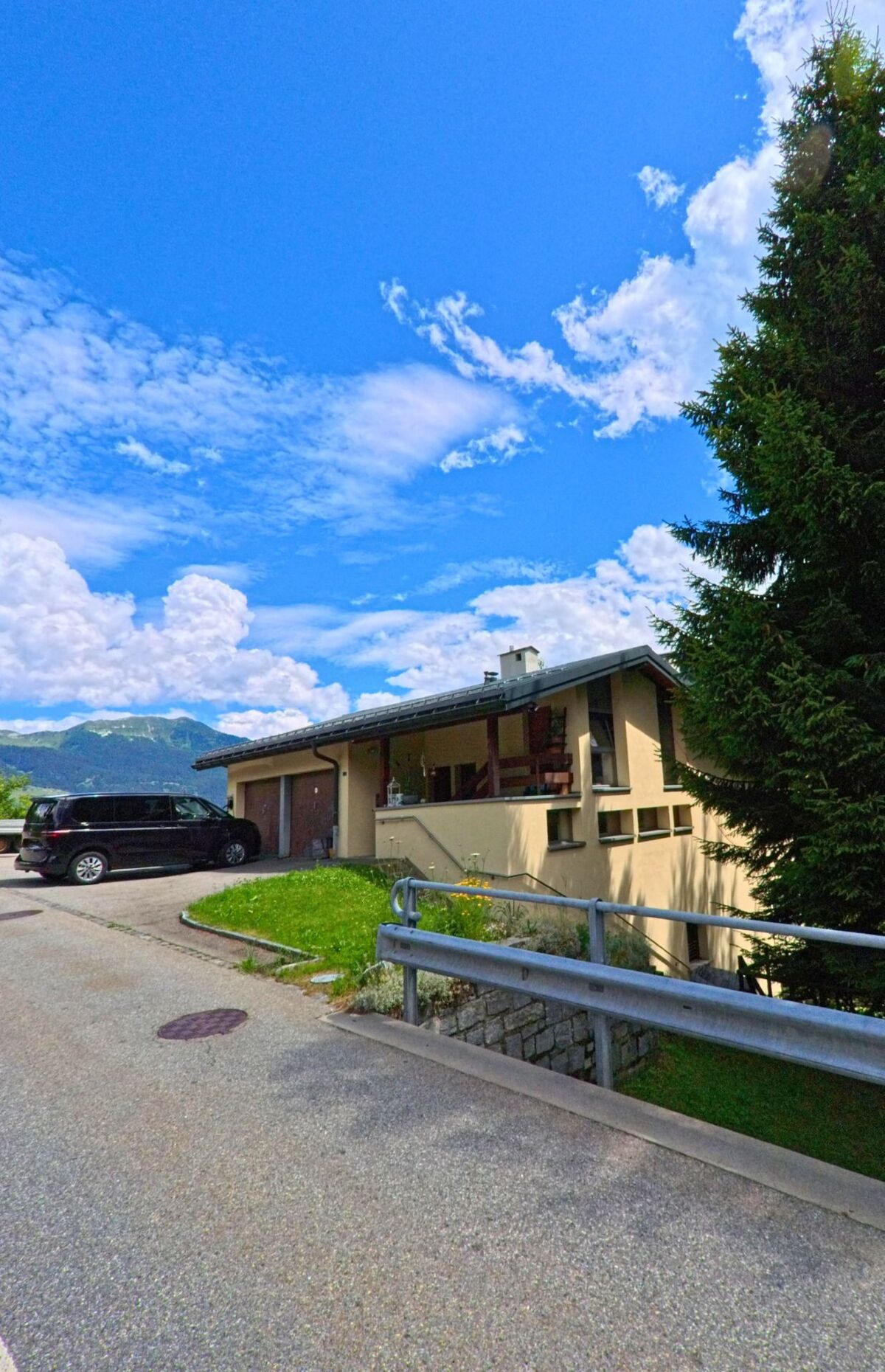 Ein modernes Einfamilienhaus mit Garagenzufahrt und vorgelagerter Tanne steht in Dalpe (6774, Kanton Tessin) an einer ruhigen Strasse mit Blick auf die Berge.