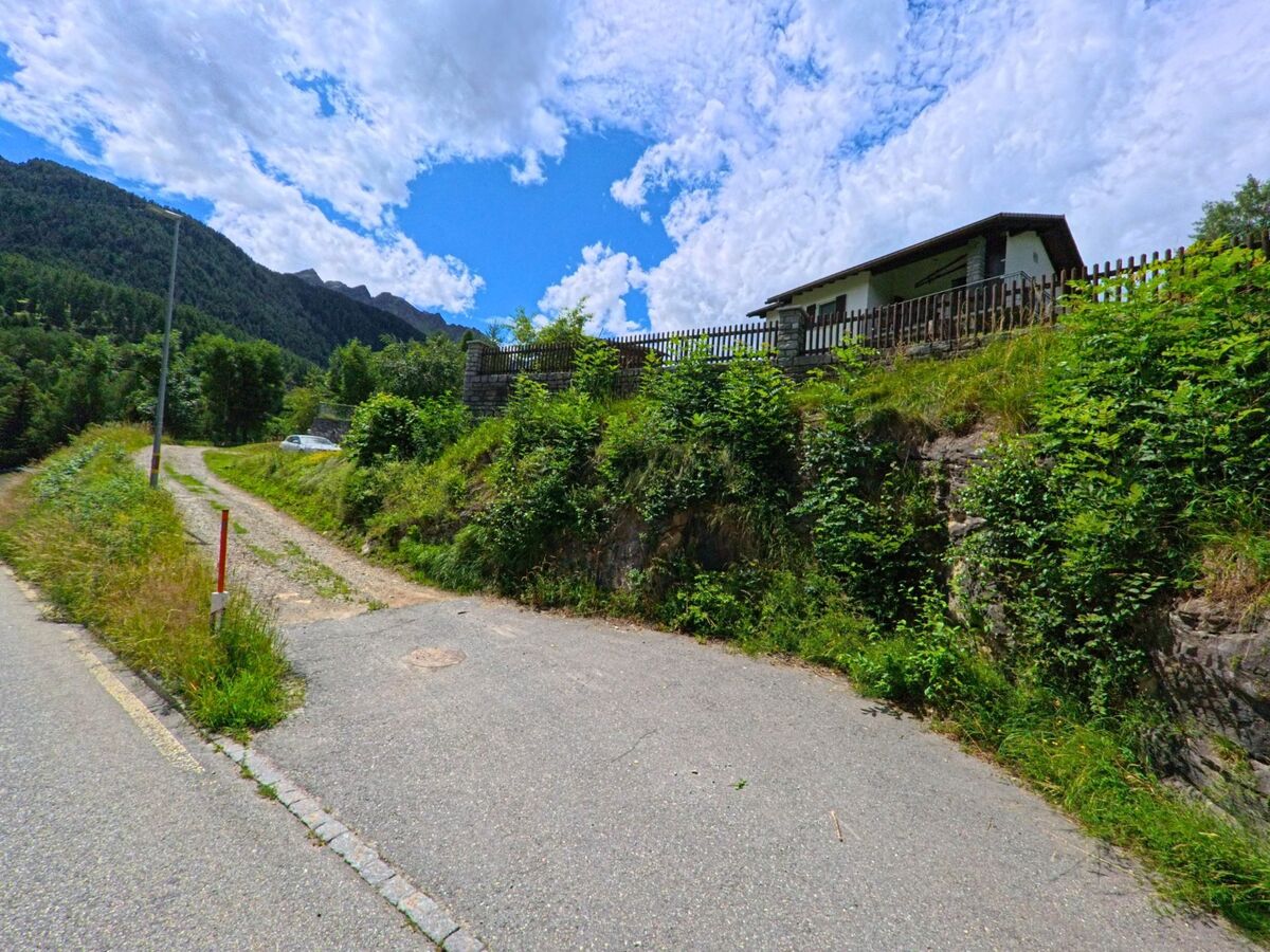 Ein schmaler Weg fuehrt in Dalpe (6774, Kanton Tessin) an einem Hang mit dichter Vegetation vorbei zu einem Haus mit Holzzaun oberhalb der Strasse.