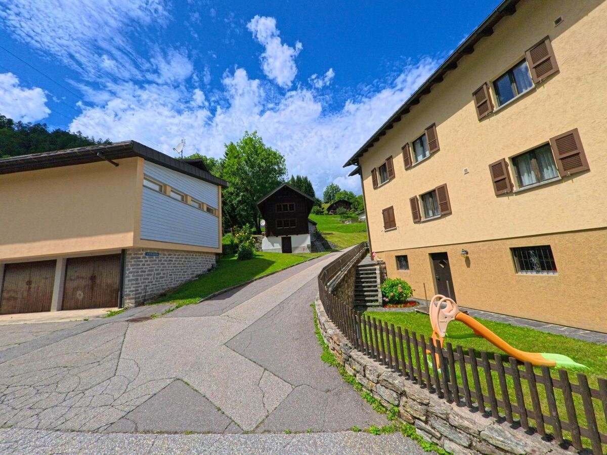 Eine schmale Anwohnerstrasse mit Wohnhaeusern und Garagen fuehrt in Dalpe (6774, Kanton Tessin) bergauf zu einem dunklen Holzhaus am Hang.