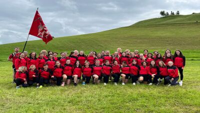 Die Mitglieder des Damenturnvereins DTV STV Schwyz stehen in Schwyz (Kanton Schwyz) in roten Trainingsjacken auf einer grünen Wiese und posieren gemeinsam mit Vereinsfahne vor einer Hügellandschaft.