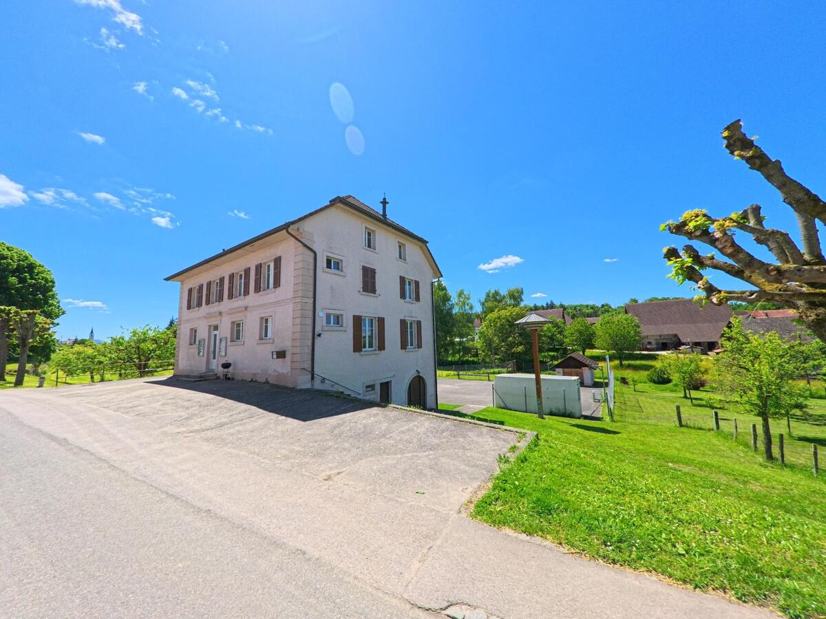 Neben einer ruhigen Strasse steht in Damphreux (2933, Kanton Jura) ein helles mehrgeschossiges Haus mit braunen Fensterläden, umgeben von gepflegten Grünflächen und Bäumen.