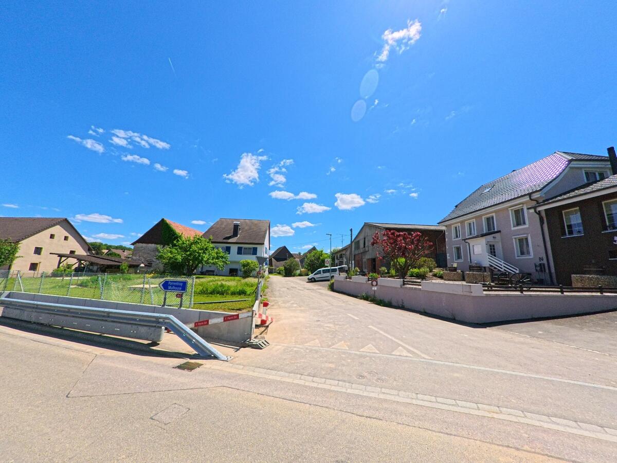 An einer Seitenstrasse in Damphreux (2933, Kanton Jura) stehen mehrere Wohn- und Landwirtschaftsgebäude neben eingezäunten Grünflächen unter einem klaren blauen Himmel.