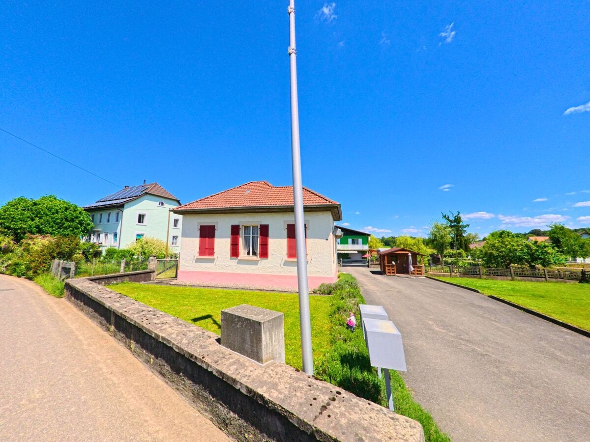 In Damphreux (2933, Kanton Jura) steht ein kleines Wohnhaus mit roten Fensterlaeden und Ziegeldach an einer Einfahrt, umgeben von gepflegten Gruenflaechen.