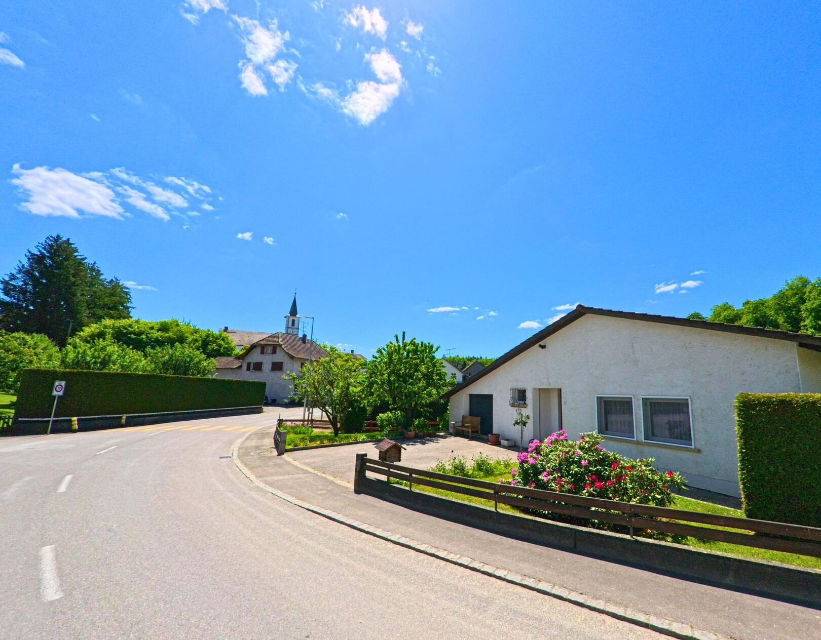 In Damphreux (2933, Kanton Jura) fuehrt eine Kurve an einem flachen Wohnhaus mit Blumengarten vorbei, im Hintergrund ist der Kirchturm des Dorfes sichtbar.