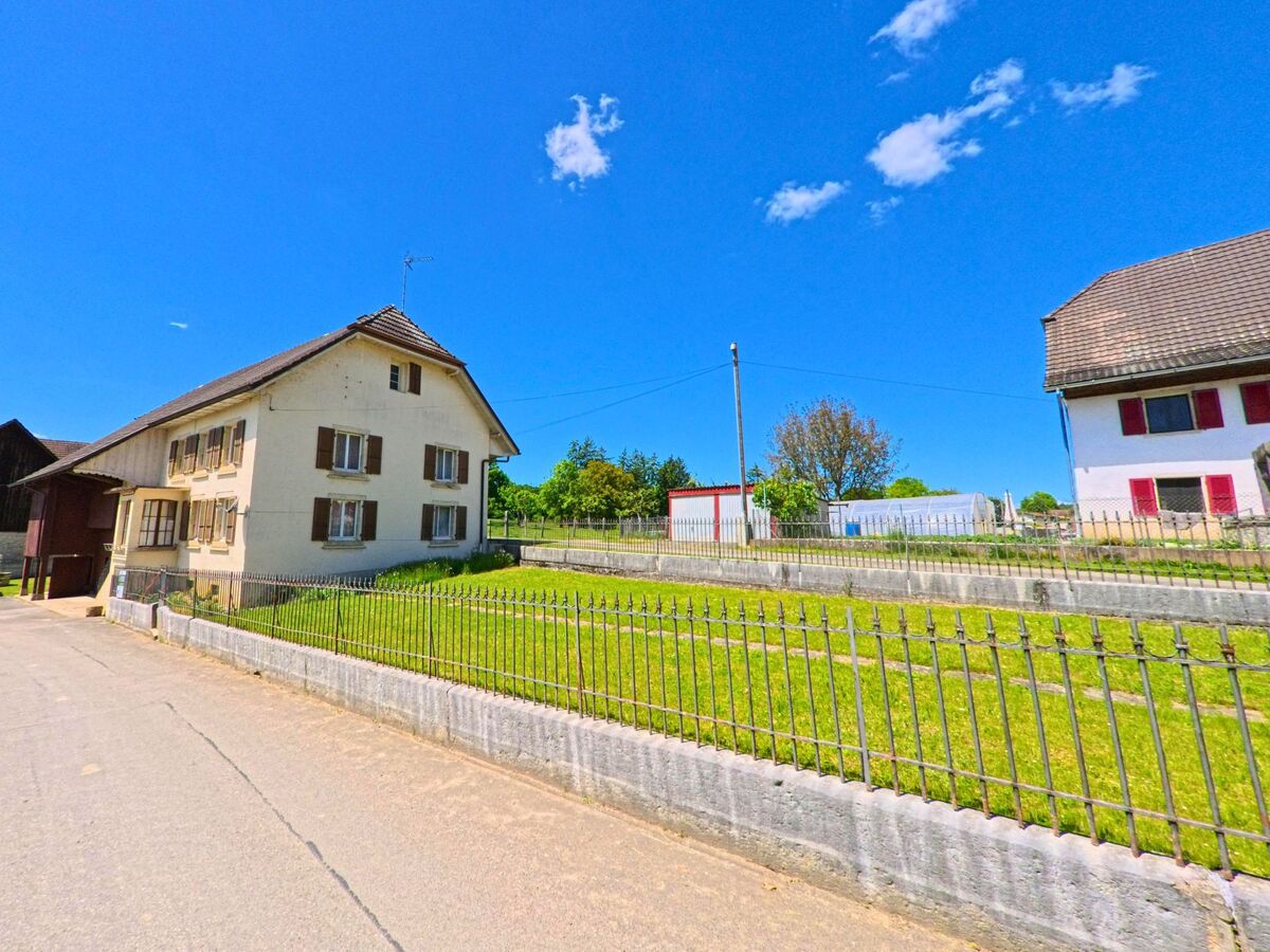 In Damphreux (PLZ 2933, Kanton Jura) steht ein Wohnhaus mit braunen Fensterlaeden an einer leicht ansteigenden Strasse, begrenzt durch einen Metallzaun und Treppenrasen.