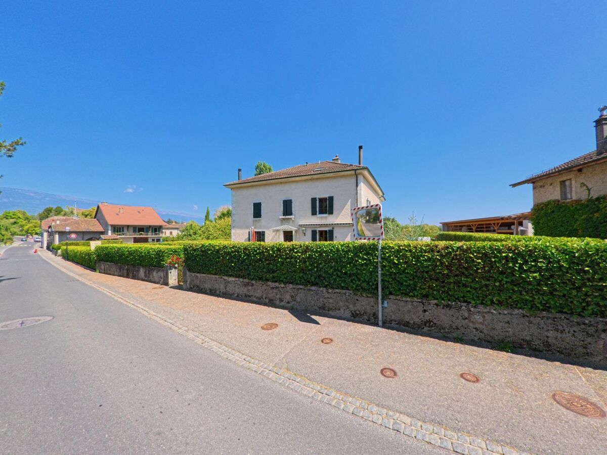Ein helles Wohnhaus mit schwarzen Fensterläden und gepflegter Hecke steht an einer Strasse in Dardagny (1283, Kanton Genf).