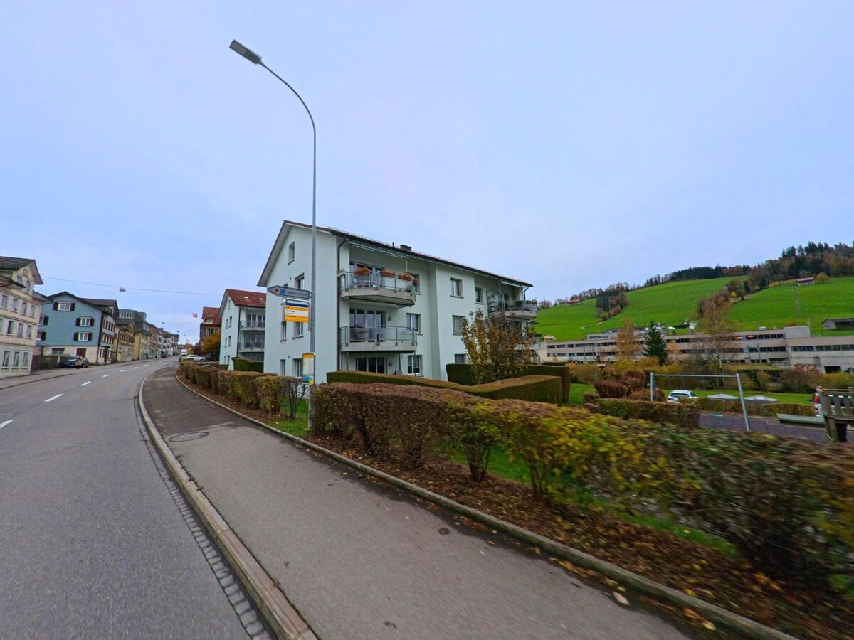 Mehrfamilienhaeuser mit Balkonen reihen sich entlang einer Hauptstrasse in Degersheim (9113, Kanton St. Gallen) vor einer offenen Huegellandschaft.