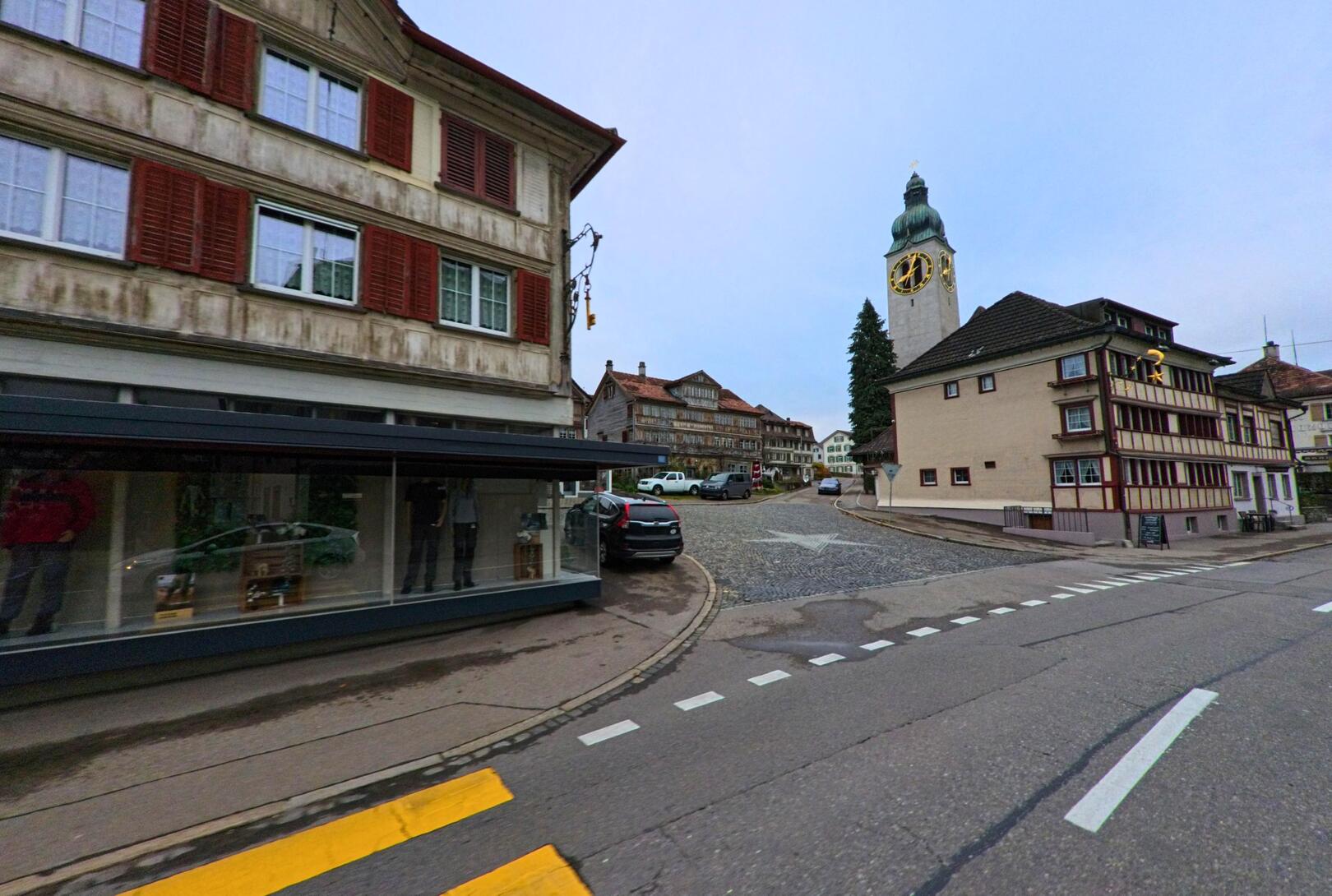 An einer gepflasterten Strassenkreuzung prägen historische Wohnhaeuser und der markante Kirchturm das Ortszentrum von Degersheim (9113, Kanton St. Gallen).