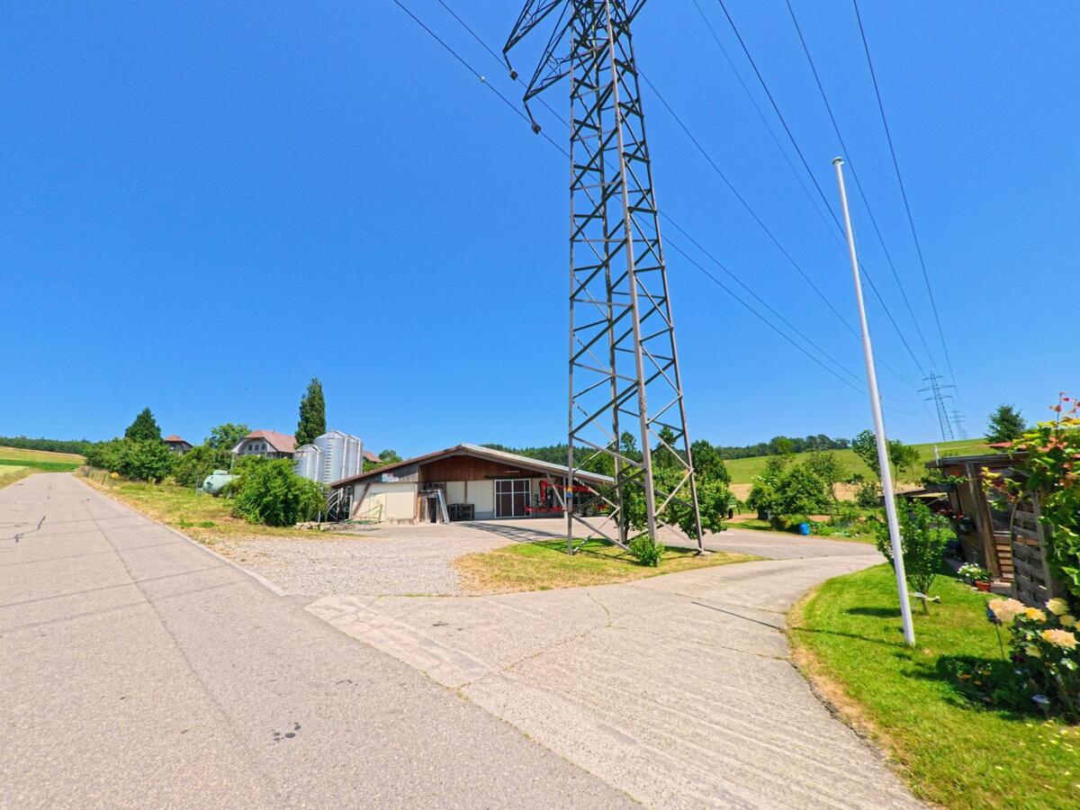 In Deisswil_bei_Münchenbuchsee (3053, Kanton Bern) führt eine geteerte Strasse an einem landwirtschaftlichen Gebäude vorbei, während ein hoher Strommast über den offenen Feldern steht.