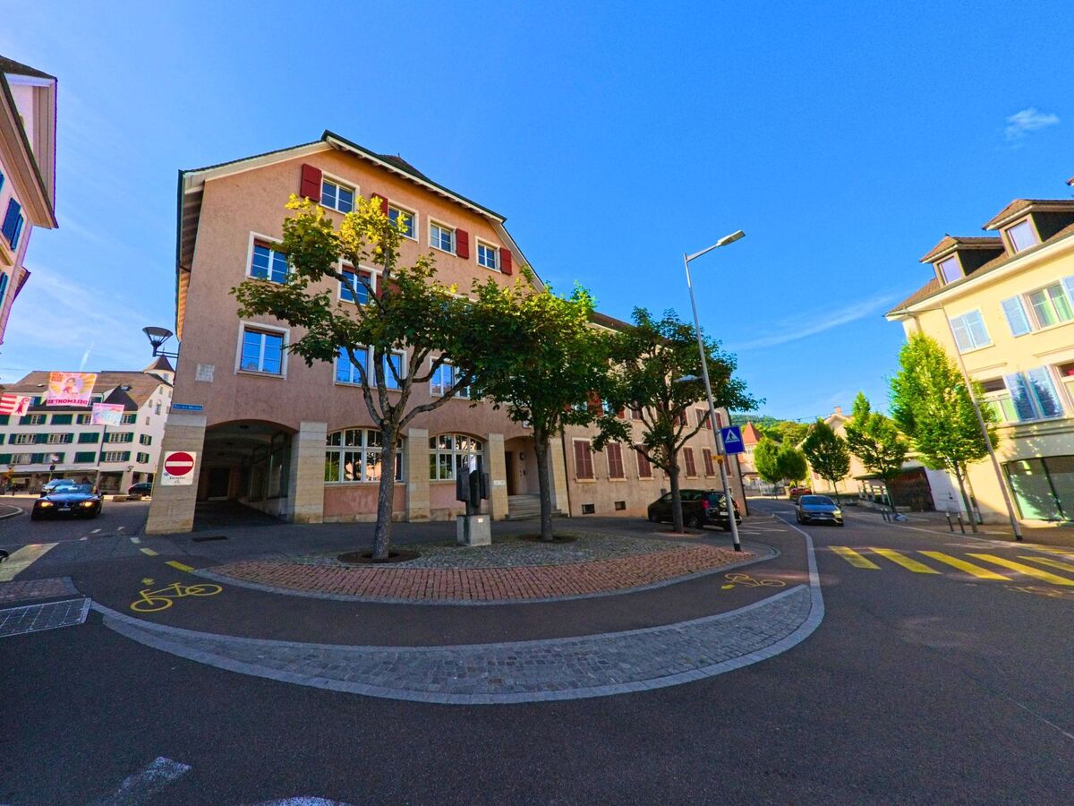 Im Zentrum von Delémont (2800, Kanton Jura) trifft eine schmale Altstadtstrasse mit Veloweg auf einen kleinen Platz, umgeben von historischen Gebäuden und Bäumen.