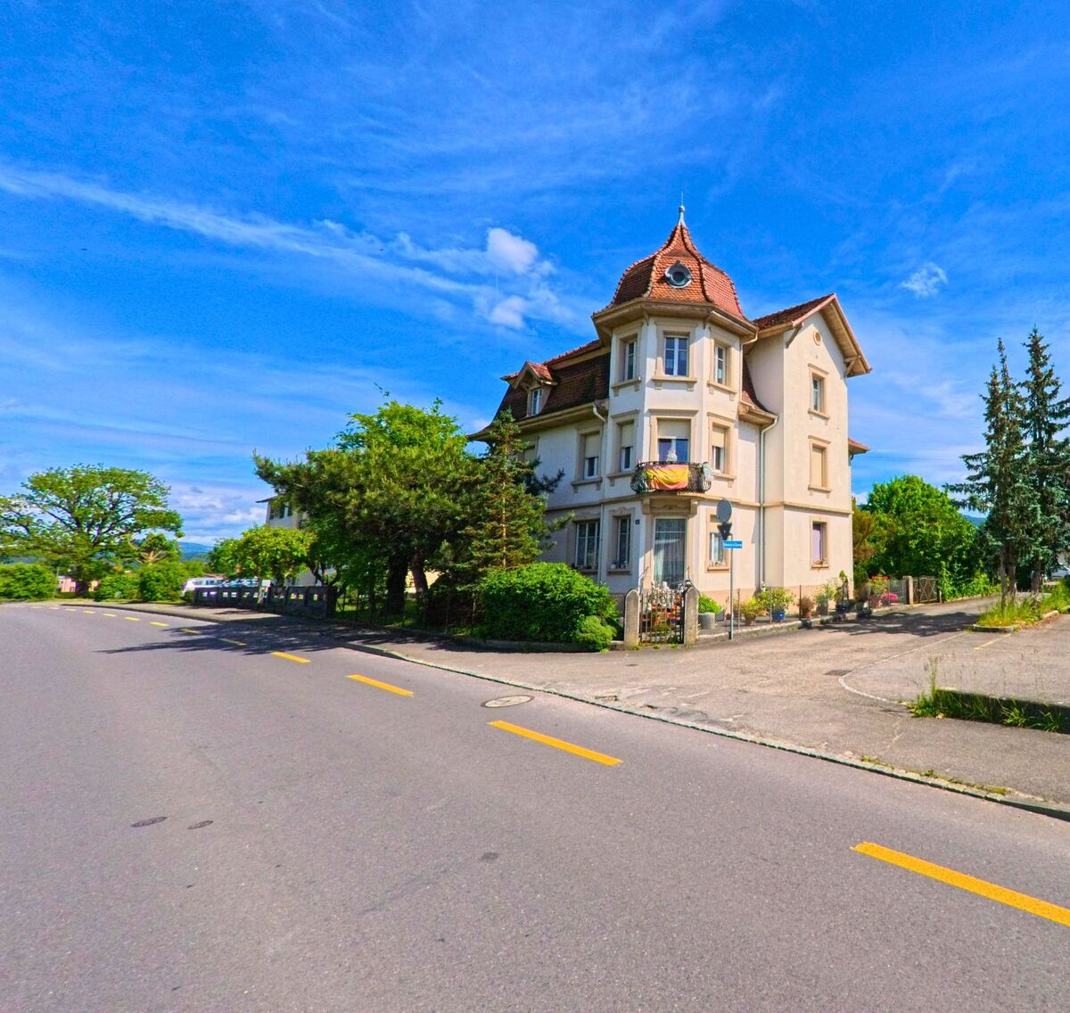 In Delémont (2800, Kanton Jura) steht ein markantes Eckhaus mit Türmchen und Ziegeldach an einer ruhigen Strasse mit angrenzendem Parkplatz.
