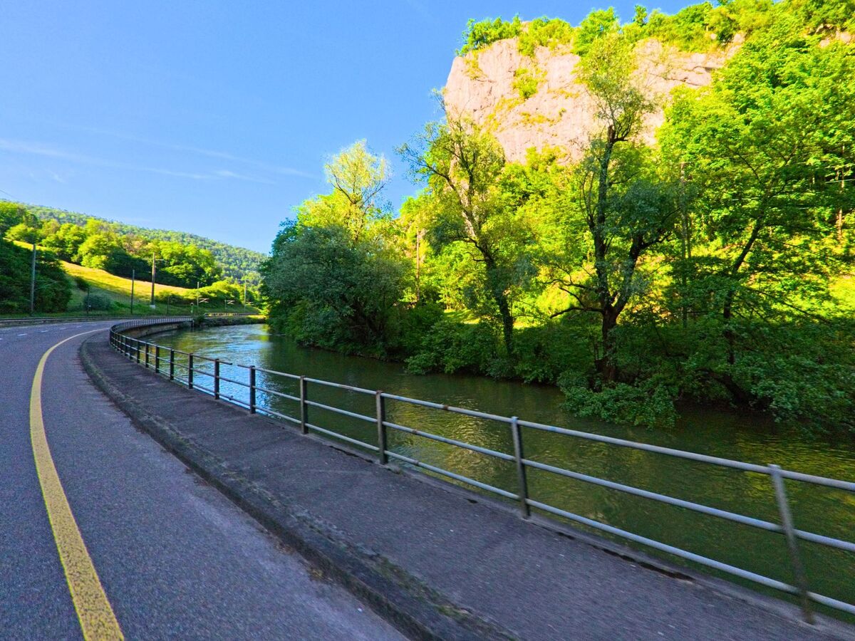 Entlang eines kurvigen Strassenabschnitts in Delémont (2800, Kanton Jura) fliesst ein Fluss am Fusse einer bewaldeten Felswand unter klarem Himmel vorbei.