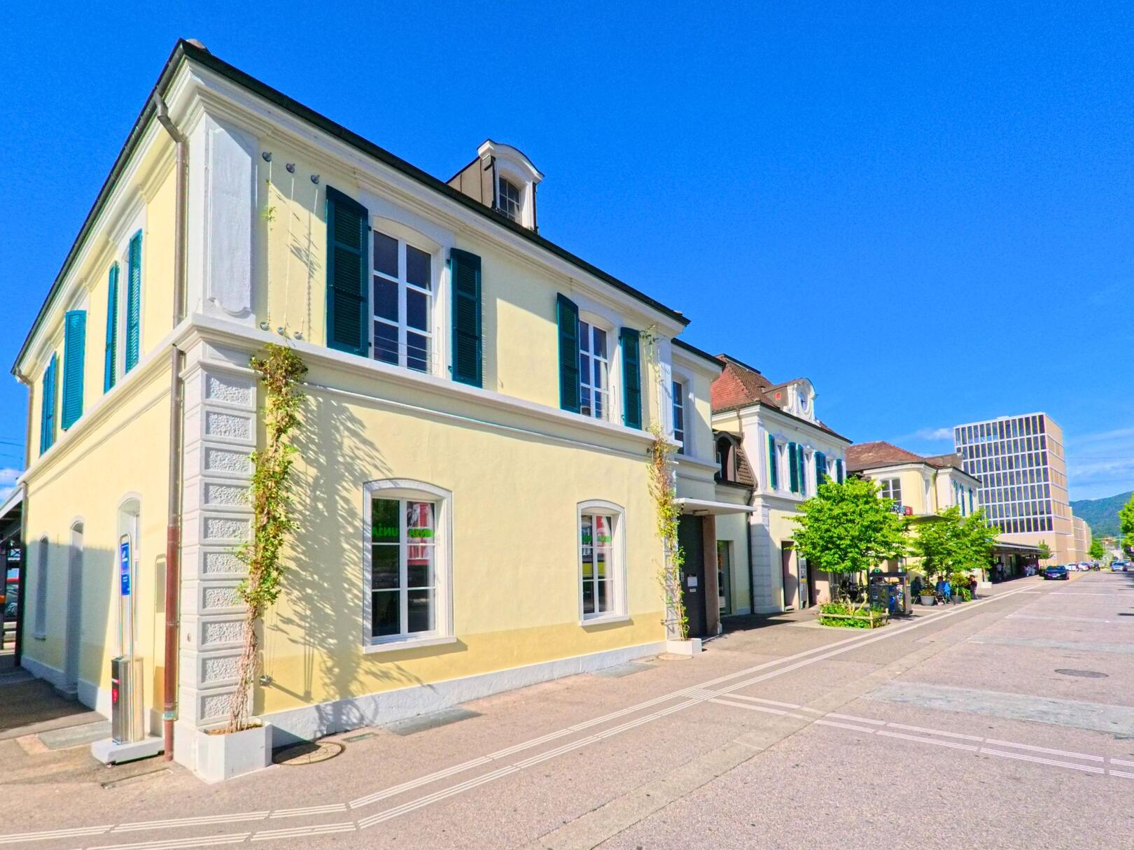 In Delémont (2800, Kanton Jura) steht ein hell gestrichenes Gebäude mit grünen Fensterläden direkt gegenüber dem Bahnhof, eingebettet in eine belebte Strassenszene.