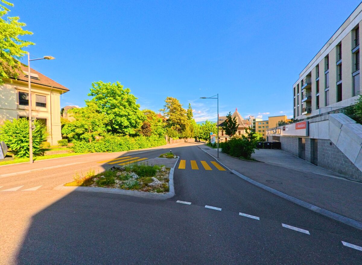 Eine verkehrsberuhigte Strasse mit Fussgängerstreifen und bepflanzter Mittelinsel führt in Delémont (2800, Kanton Jura) durch ein gemischt genutztes Quartier mit moderner und historischer Bebauung.
