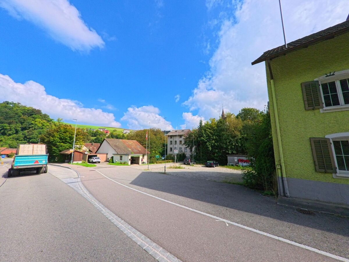 Zwischen Wohnhäusern und dem Gemeindezentrum von Densbüren (5026, Kanton Aargau) verläuft eine ruhige Strasse mit Blick auf die umliegenden Hügel.