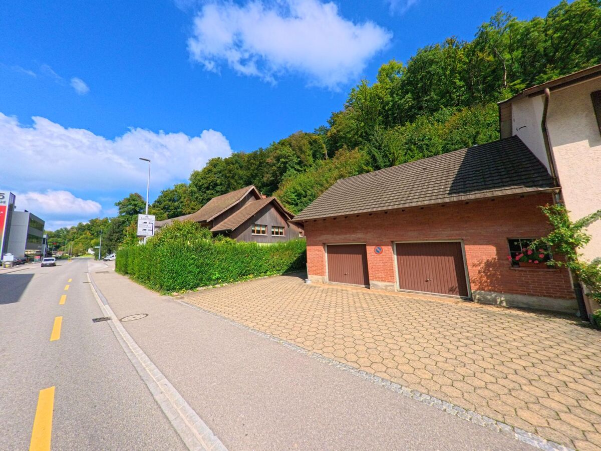 Entlang der Strasse in Densbüren (5026, Kanton Aargau) stehen Wohnhäuser und Garagen mit gepflegten Vorplätzen am Fuss eines bewaldeten Hangs.