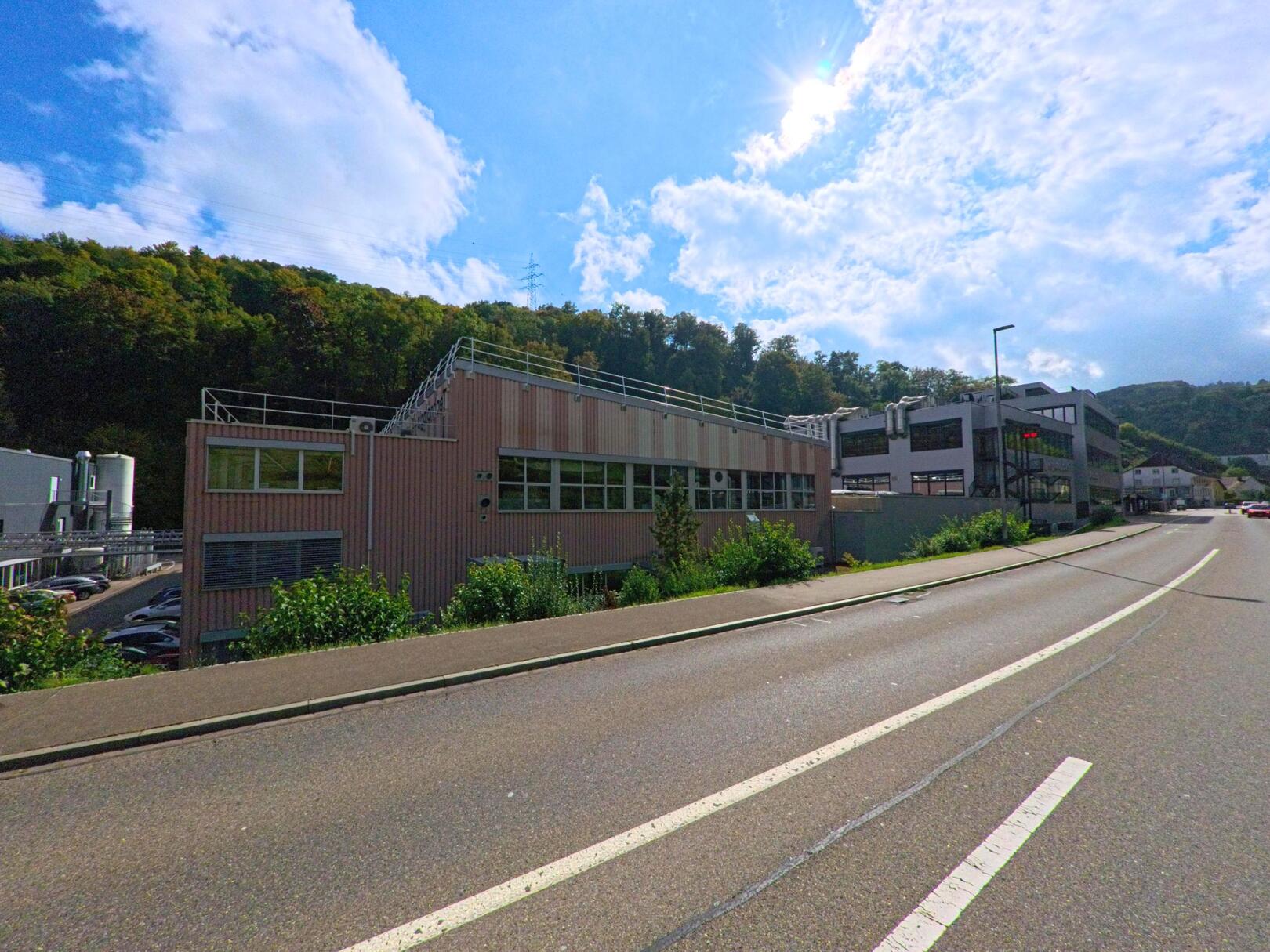 Entlang der Hauptstrasse in Densbüren (5026, Kanton Aargau) stehen moderne Gewerbebauten vor bewaldeten Hügeln unter einem hellen Himmel.