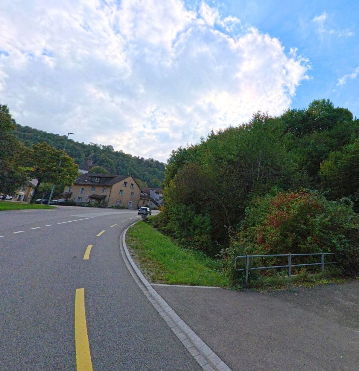 Eine sanft geschwungene Strasse in Densbüren (5026, Kanton Aargau) führt an Wohnhäusern und dichtem Grün vorbei in Richtung des Dorfzentrums.