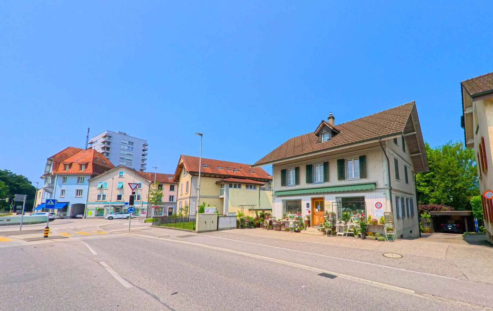 In Derendingen (4552, Kanton Solothurn) befindet sich an einer belebten Kreuzung ein Geschäftshaus mit Blumenladen, umgeben von Wohnhäusern und einem Hochhaus im Hintergrund.