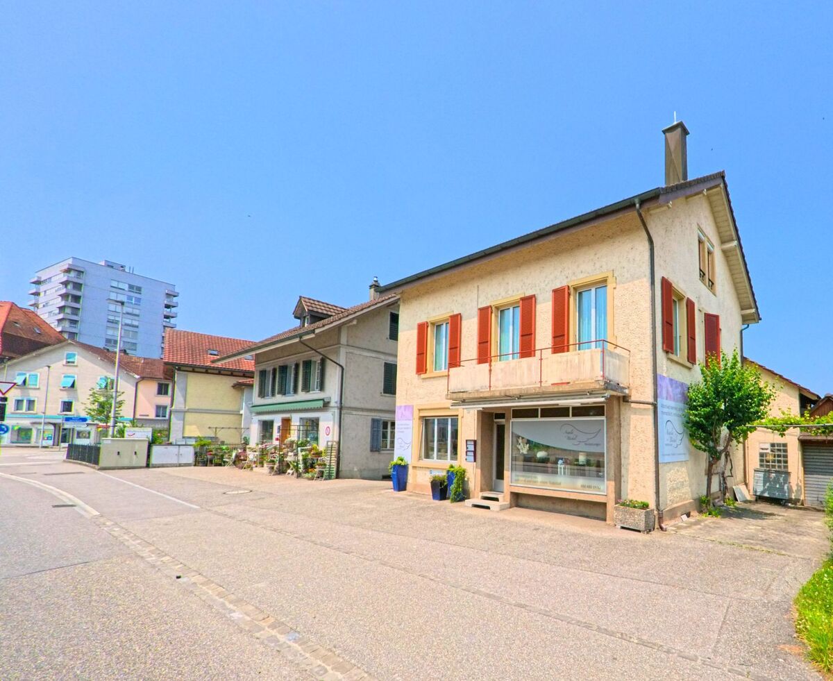 In Derendingen (4552, Kanton Solothurn) stehen Geschaeftshaeuser mit roten Fensterlaeden und Ladengeschaeften entlang einer Hauptstrasse, im Hintergrund ist ein Hochhaus sichtbar.