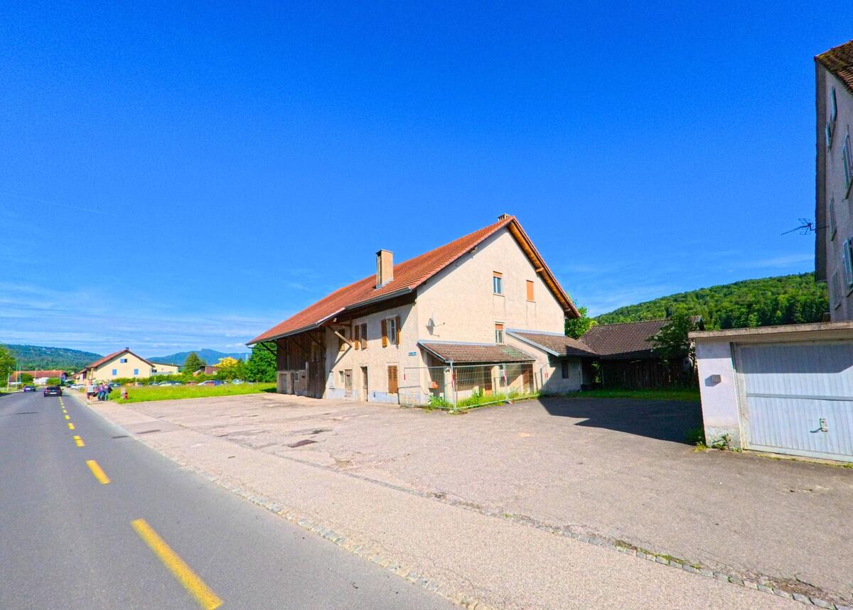 In Develier (2802, Kanton Jura) führt die Straße an einem älteren Gebäude und einer Garage entlang, während die Hügel im Hintergrund zu sehen sind.