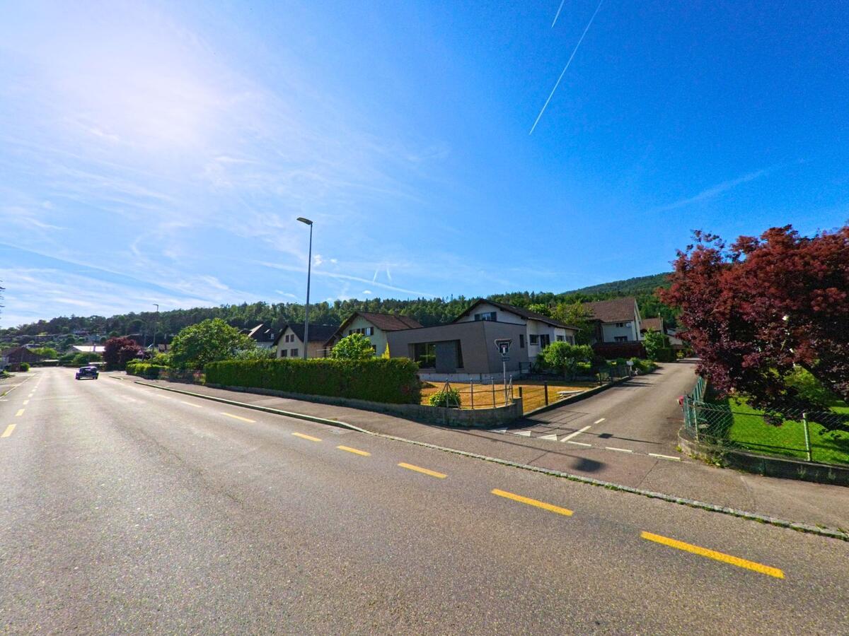 In Develier (2802, Kanton Jura) führt eine sonnige Straße an gepflegten Häusern und grünen Bäumen vorbei, mit Hügeln im Hintergrund.